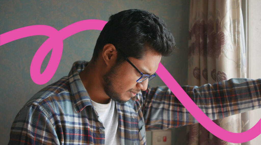 Man with dark hair and glasses wearing a plaid shirt looks down, standing indoors with a pink decorative swoosh overlaying the image.