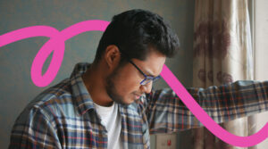 Man with dark hair and glasses wearing a plaid shirt looks down, standing indoors with a pink decorative swoosh overlaying the image.
