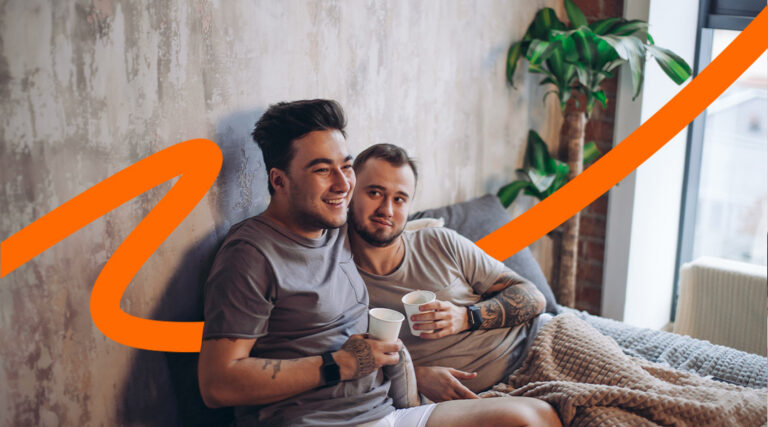 Two men sit on a sofa in a cozy living room, smiling and holding disposable cups, with an orange decorative line crossing the image.
